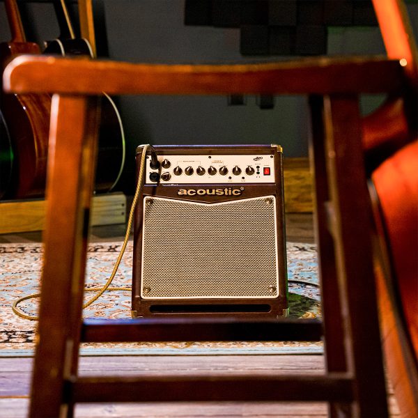 A20 20W acoustic amp behind the stool with guitar leaning to it, with acoustic guitar in the background