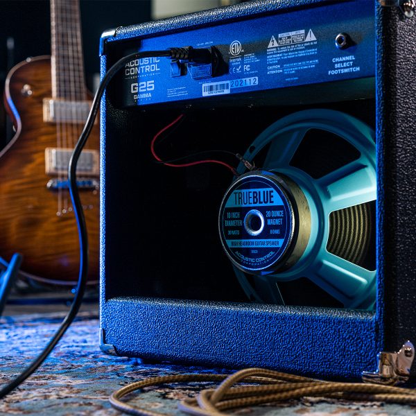 The open back of a GAMMA G25 guitar amp exposes a bright blue TRUE BLUE speaker inside. To the side is an electric guitar.