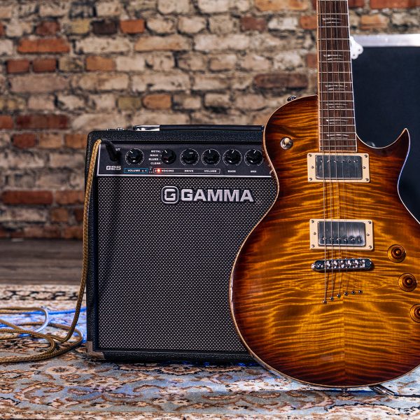 GAMMA G25 electric guitar amp plugged in in front of a brick wall. An electric guitar leans against the amp.