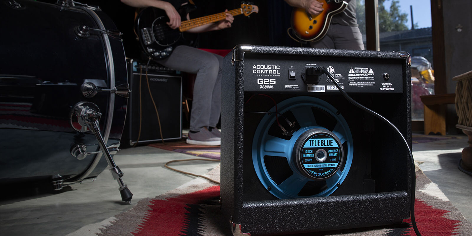 The open back of a GAMMA G25 guitar amp exposes a TRUE BLUE speaker inside. In front of the amp are guitar players and a drum set.