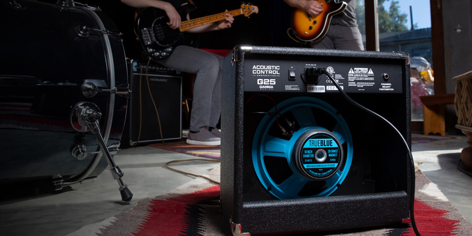 The open back of a GAMMA G25 guitar amp exposes a TRUE BLUE speaker inside. In front of the amp are guitar players and a drum set.